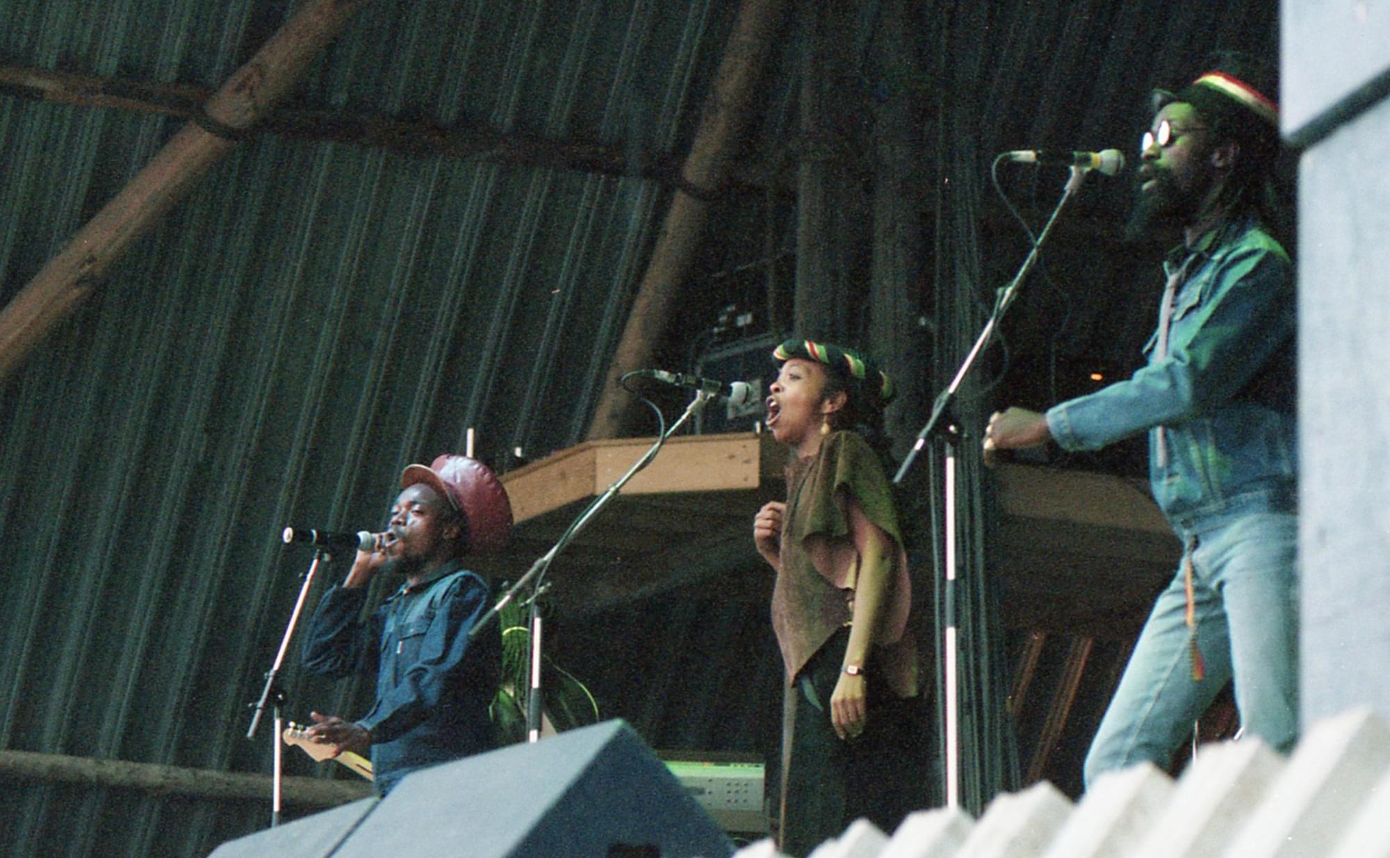 Black Uhuru perform on the Pyramid Stage. Three performers stand on a stage, each with a microphone stand in front of them. Closest to the camera stands a man wearing a denim jacket, jeans, a black hat and sunglasses. To his right a woman is singing into the microphone wearing a brown short sleeved top, a black skirt or trousers and a green, red, yellow and black head band. To her right a man with a red hat and blue shirt holds the microphone stand and sings into it.
