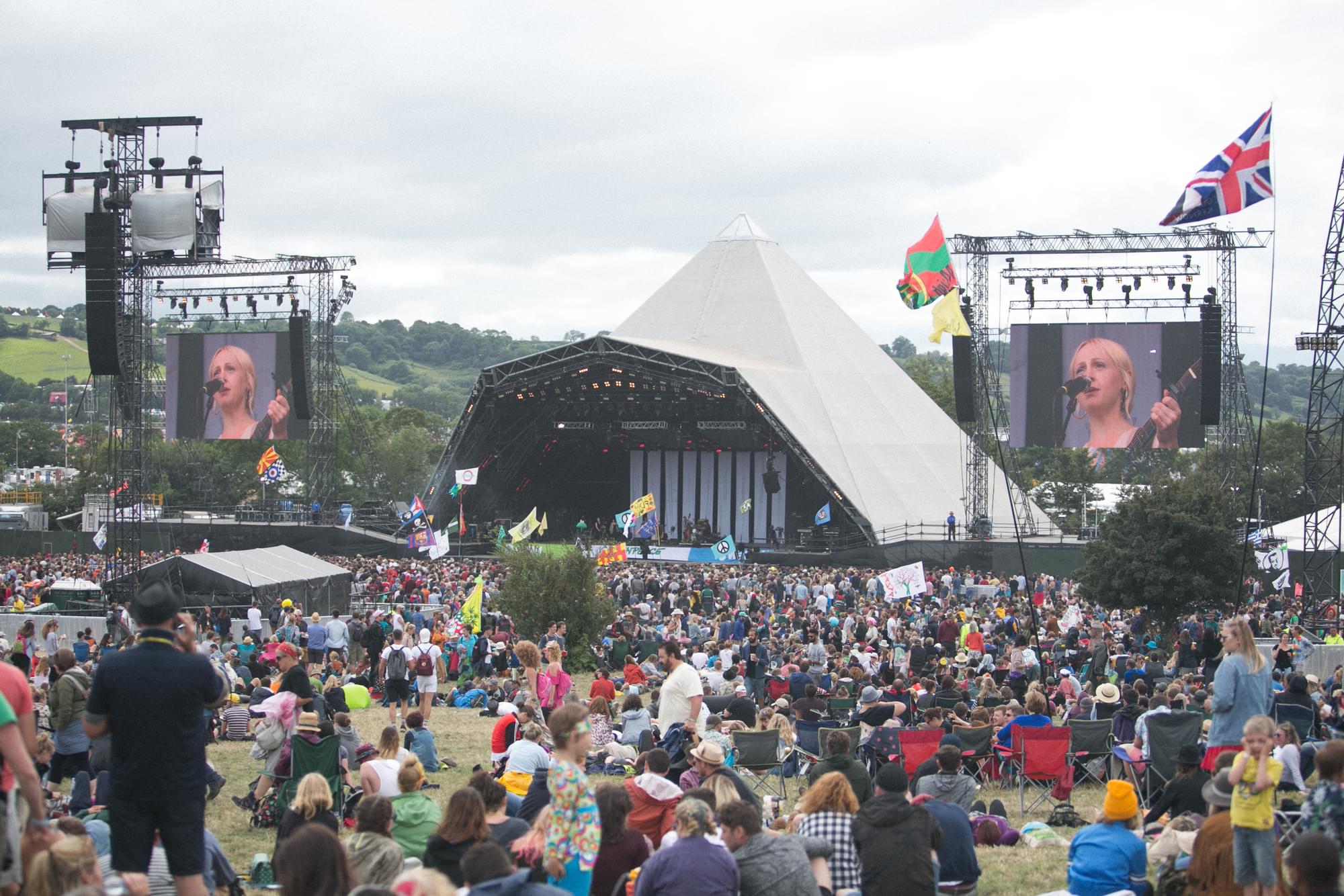 A photograph taken during the day at Glastonbury festival depicting a performance by Laura Marling. The image shows a pyramid shaped stage in a field with a large crowd gathered in front if it. Colourful flags and banners are held above the crowd. Two large video screens are installed on either side of the stage and showing on both screens is an upclose shot of Laura Marling performing in front of a microphone stand and holding a guitar.