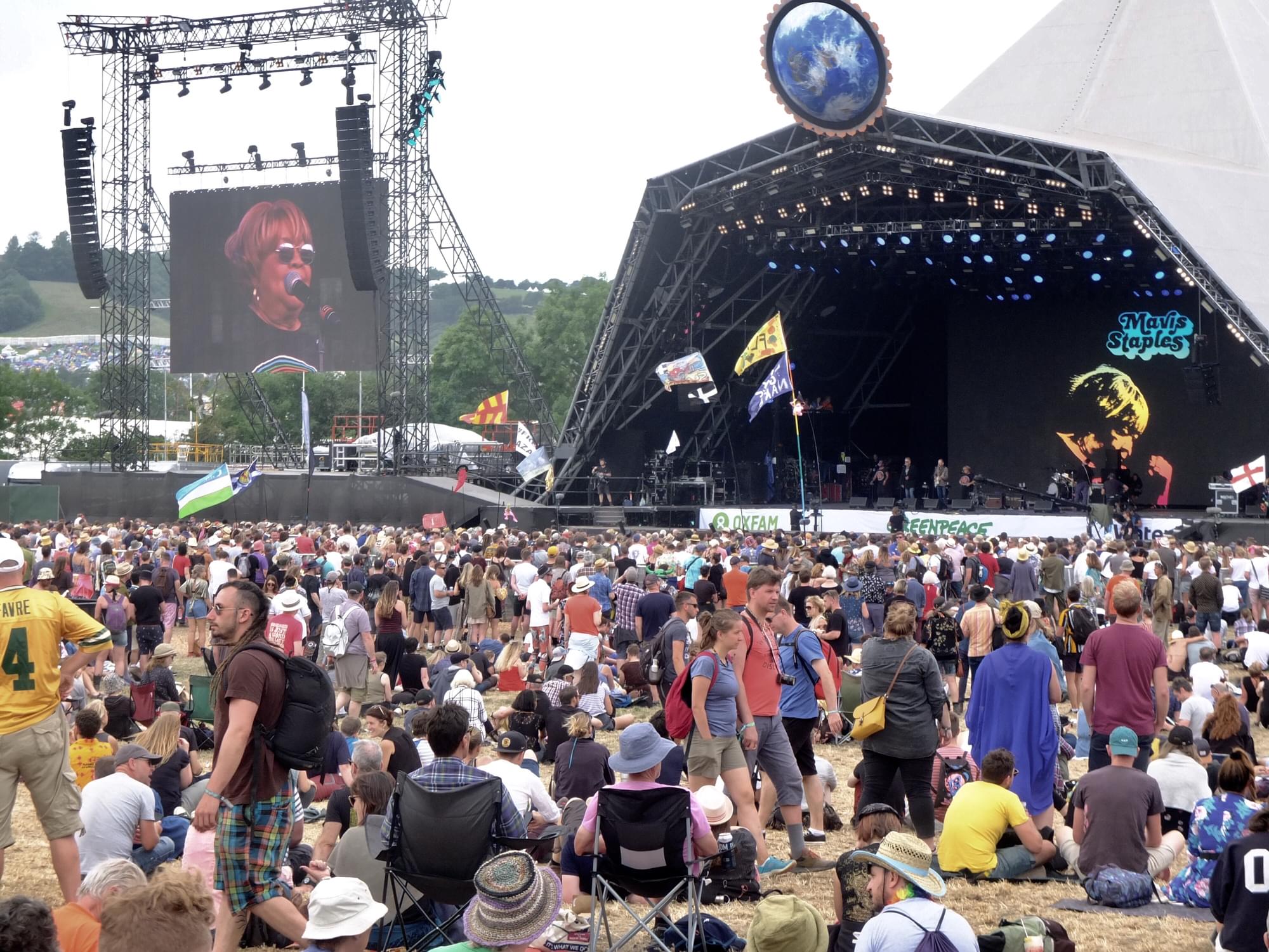 A photograph taken at Glastonbury Festival during the day time depicting a performance on a stage by Mavis Staples. A large crowd stand in front of a stage. A group of performers are visible on the stage standing in front of a black back drop that says Mavis Staples in blue letters and shows a side view image of Mavis Staples face in orange and yellow. To the left of the stage a large video screen is installed showing an upclose shot of Mavis Staples standing in front of a microphone.