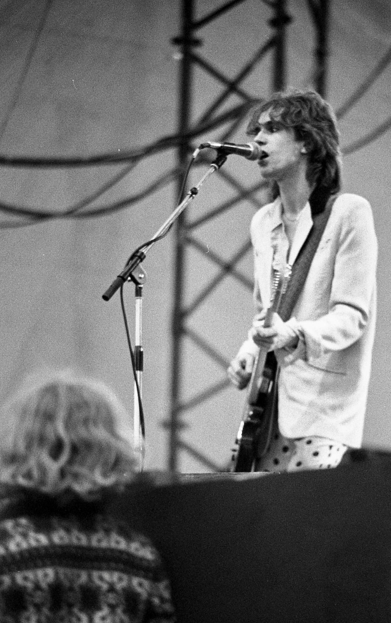 A black and white photograph of the guitarist from the Only Ones playing on stage and singing into a microphone. In front of the stage, a person wearing a patterned jumper looks up towards the musician, with their back to the camera.