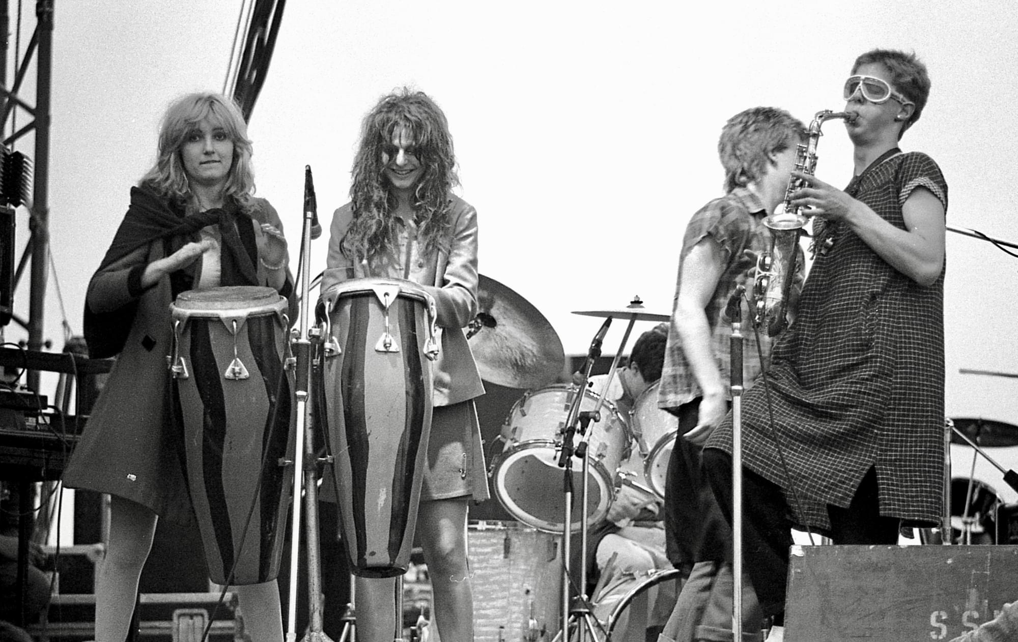 A black and white photograph of The Slits performing on stage. Two female members of the band stand behind bongo drums. A third female is performing on a saxophone. The fourth person is looking away from the camera, towards the left side of the stage. A drum kit is visible in the background.