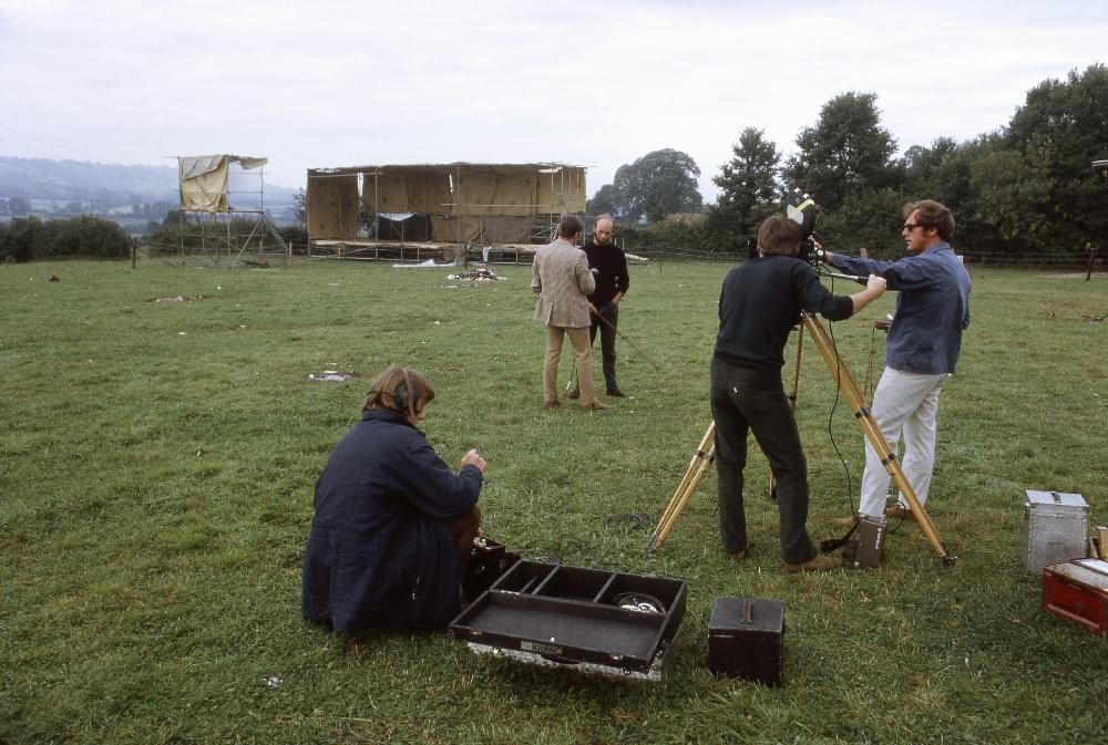 A photograph taken at Glastonbury during the daytime in 1970. A crew of three people are setting up a tripod camera and pointing it away from the camera towards a field. In the field stands Michael Eavis, dressed all in black, being interviewed by another man in a brown suit. Behind Michael a temporary stage is visible at the other end of the field.